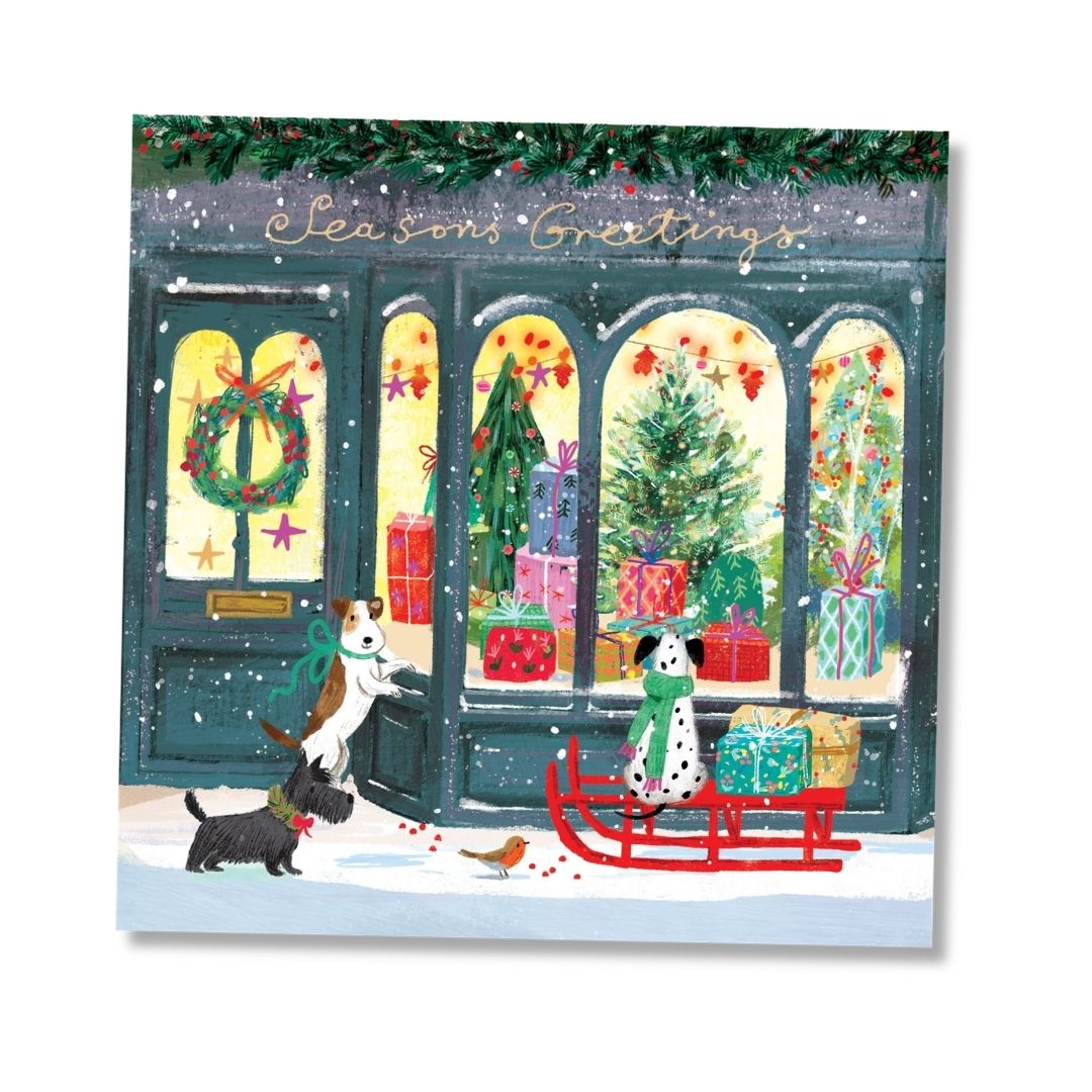 A square Christmas greeting card featuring an illustrated storefront scene with snow, garlands, wrapped gifts, and two dogs near a red sled. The card is shown against a clean white background with a soft drop shadow.