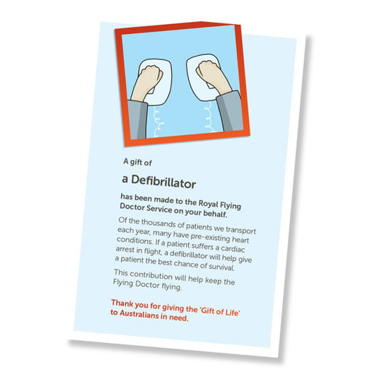 A card for a donation of an defibrillator, shown at an angle on a white background. The card has a pale blue field, a small illustrated header with hands on defibrillator paddles, and a red-orange border, and text stating it is a gift of a defribillator and a that it is a donation to the Royal Flying Doctor Service.
