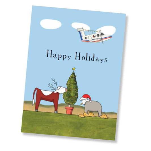 A holiday greeting card shown at a slight angle on a white background. The card features a light blue sky background with the words 'Happy Holidays' in black, an illustrated airplane, a green Christmas tree, a red-and-white cow, and a gray sheep wearing a Santa hat.