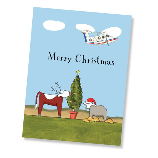 A Christmas greeting card shown at a slight angle on a white background. The card features a light blue sky background with the words Merry Christmas' in black, an illustrated  RFDS plane, a green Christmas tree, a red-and-white cow, and a gray sheep wearing a Santa hat.
