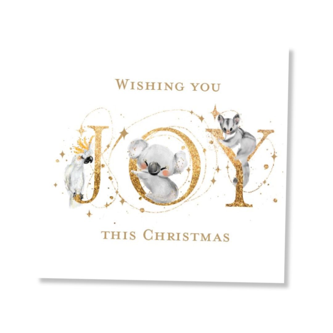 A square white Christmas greeting card featuring the word 'JOY' in large gold glitter letters, decorated with a cockatoo, koala and ring-tail possum nestled within the letters. The text reads 'Wishing you JOY THIS CHRISTMAS' in gold.